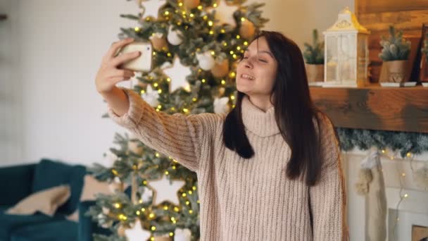 Happy young lady is talking to friends online making video call with smartphone on Christmas day standing near fir-tree and decorated mantel at home. - Imágenes, Vídeo