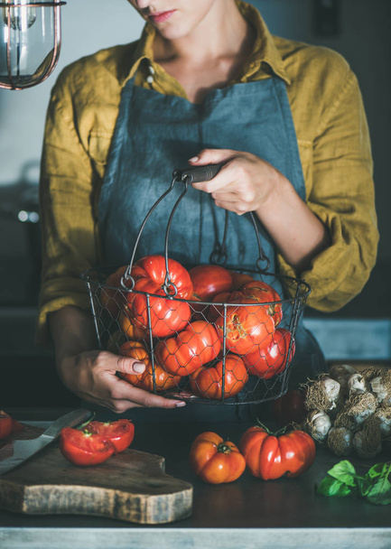 Woman in grey linen apron holding basket with heirloom tomatoes and cooking tomato sauce, canned tomatoes or pasta with basil and garlic at kitchen counter. Healthy cooking, slow food or comfort food - Photo, Image