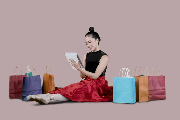 Молодая балерина с помощью цифровой таблетки для покупок в Интернете, сидя рядом с бумажными пакетами в студии
 - Фото, изображение