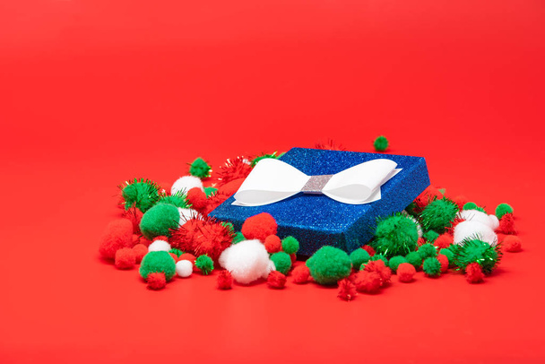 Blauwe doos van de gift van Kerstmis met een boog omringd door rode, groene en witte pompom. Geïsoleerd op rode achtergrond. - Foto, afbeelding