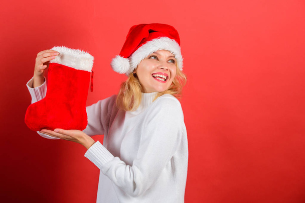 Girl cheerful face got gift in christmas sock. Christmas stocking concept. Traditional winter holiday. Check contents of christmas stocking. Woman in santa hat hold christmas gift red background - Photo, image
