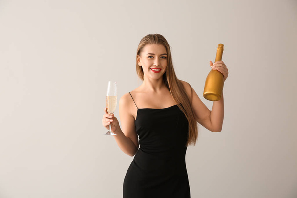 Beautiful young woman with glass and bottle of champagne on light background - 写真・画像