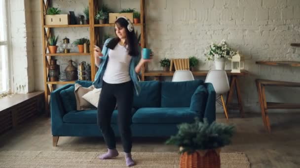 Attractive brunette is having fun at home listening to music through headphones and dancing holding smartphone and cup. Modern technology and people concept. - Séquence, vidéo