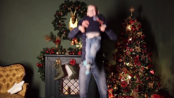 Father and son in festive Santa s hats playing and twisting near the decorated Christmas tree. Father is twisting around with his son on hands. Kissing his son. New Year concept. - Footage, Video
