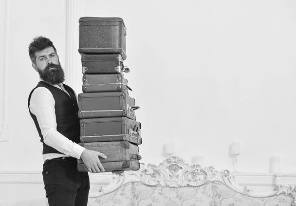 Man with beard and mustache wearing classic suit delivers luggage, luxury white interior background. Macho, elegant porter carries heavy pile of vintage suitcases. Butler and service concept - Foto, Imagem