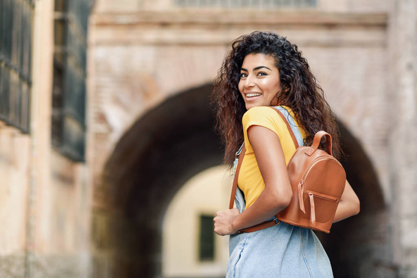 Rear view of young Arab woman with backpack outdoors. Traveler girl in casual clothes in the street. Happy female wearing yellow t-shirt and denim dress in urban background. - Foto, immagini