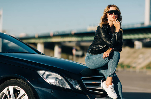 Image of young blonde woman in sunglasses sitting on hood of black car - Fotó, kép