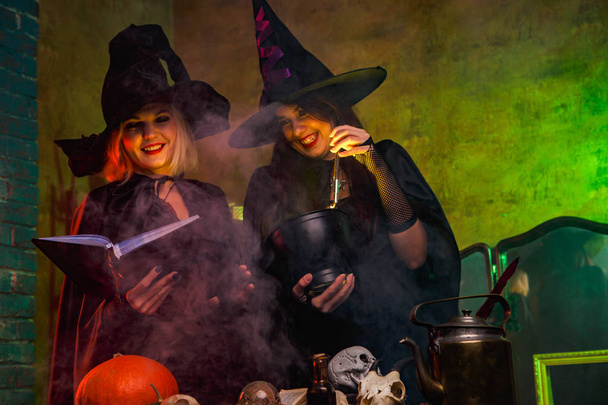 Mosolyogva két boszorkányok a fekete kalap könyvével asztalhoz a tök és a koponyákat portréja - Fotó, kép