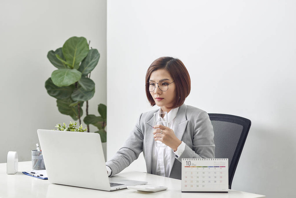 Relajada mujer de negocios madura sosteniendo un vaso de agua y trabajando en su portátil en la oficina
 - Foto, imagen