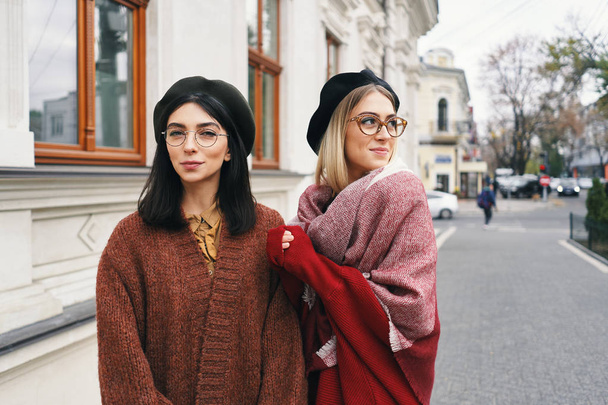 Carefree fashionable women models in elegant autumn clothes and glasses. Young fashion girls in stylish woven wool clothes and hats over urban city background, autumn portrait.  - Fotoğraf, Görsel