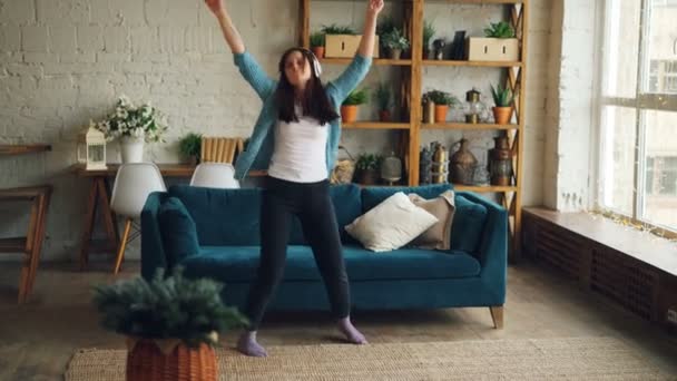 Carefree young woman is dancing at home listening to music through headphones and moving raising arms indoors in modern apartment. People and fun concept. - Záběry, video
