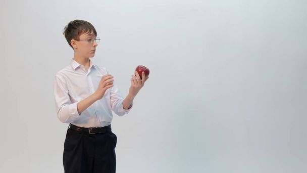 Staunend blickt der Teenager mit der Brille auf einen roten Apfel. Lustige Emotionen im Bild eines hervorragenden Studenten. Schuljunge in schwarzer Hose und weißem Hemd mit hochgekrempelten Ärmeln auf hellem Hintergrund. - Foto, Bild