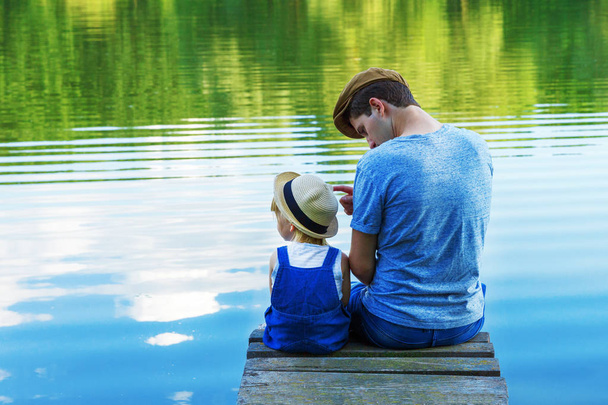 Dad and a tiny girl in dungarees relax at the river. Back view, copy space. - Foto, imagen