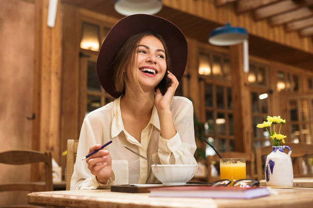 Portrait of elegant woman wearing hat talking on smartphone while sitting in cozy cafe - Fotografie, Obrázek