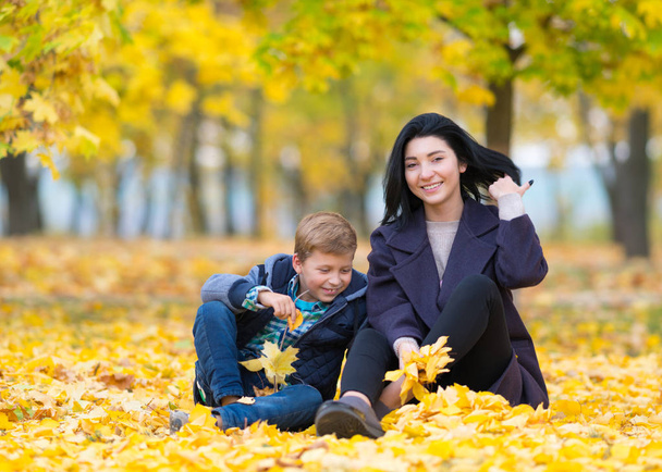 A happy, content mother and son sitting and playing in a city park amongst yellow autumn leaves. - Valokuva, kuva