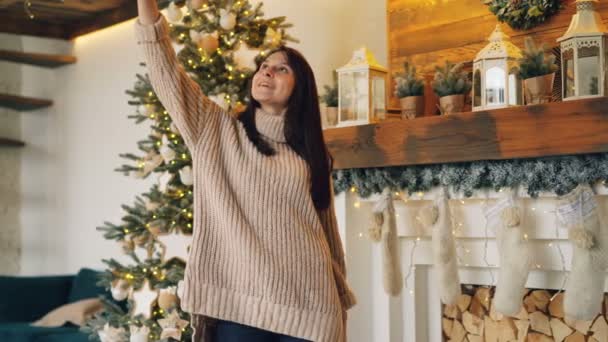 Carefree girl is taking selfie on Christmas day standing near New Year tree and fireplace holding smartphone and posing for camera. Holidays and photos concept. - Video, Çekim