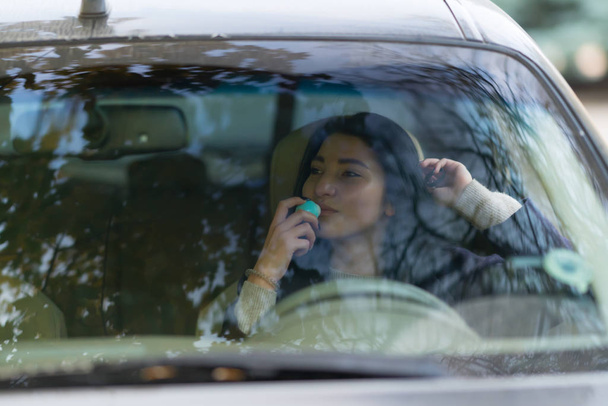 Young brunette woman behind the wheel of a car and sniffing round blue object in her hand and looking away, viewed from her front through the windshield. Portrait with copy space - Foto, Imagem
