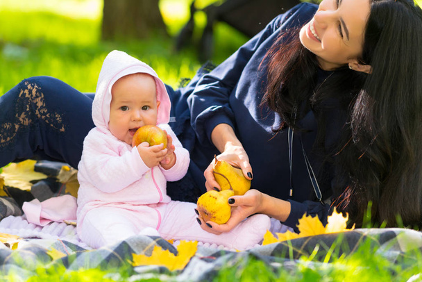 Happy young mother laughing at her baby girl as she sits on a rug on the grass in an autumn park holding a ripe golden apple - Valokuva, kuva