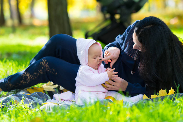 Loving young mother in a park with her baby daughter lying together on a blanket on the grass playing with colorful autumn leaves - Photo, Image