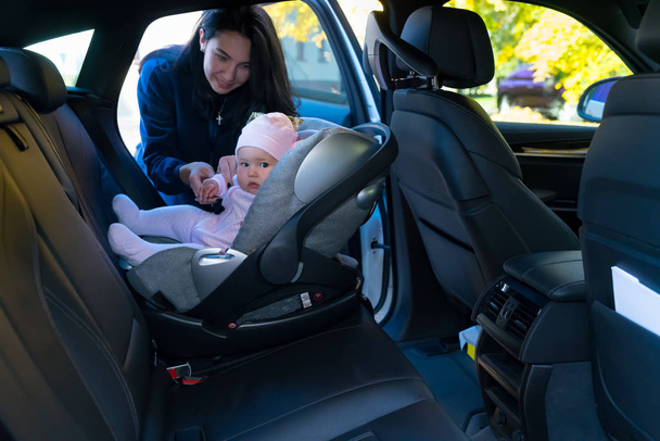 Young mother standing next to her baby sitting in baby seat backwards on backseat of the car with black interior - Photo, Image