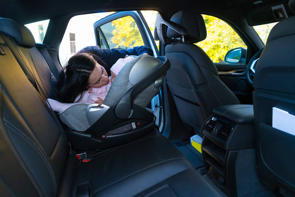 Mother kissing her baby in baby seat on the backseat of the car with black interior, viewed from other side of the vehicle with copy space - Photo, Image