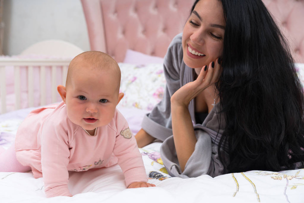 Young mother with long black hair with her young baby girl in bed, looking at her daughter and smiling. The child is dressed in pink dress, standing on all fours and looking at camera - Zdjęcie, obraz