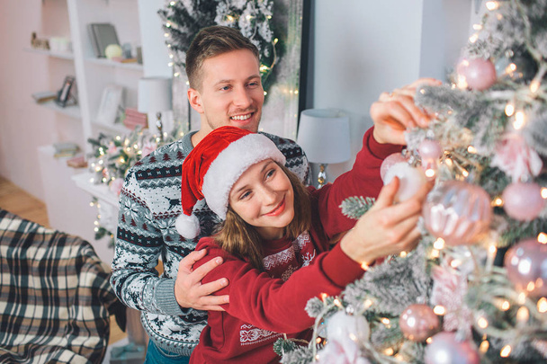 Young woman decorating Christmas tree. She touches toy on it. Young man stands behind and embrace her. They smile. - Foto, immagini