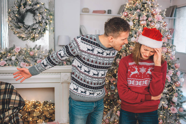 Angry young man stands close to woman and screams at her. He points left. Model is upset. She cries. They wear Christmas clothes. - Photo, Image