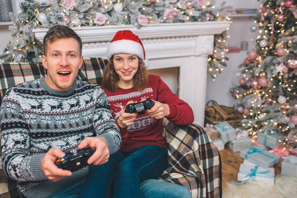 Two people sitting and playing games. Thay hold gamepads in hands. Young man and woman are excited. They push buttons and play with excitement in decorated room. - Фото, зображення