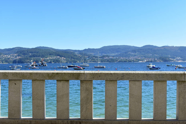 Hintergrund: Steingeländer in einer Strandpromenade. Türkises Wasser in einer Bucht, Boote und blauer Himmel. sonniger tag, galicien, rias baixas, spanien. - Foto, Bild