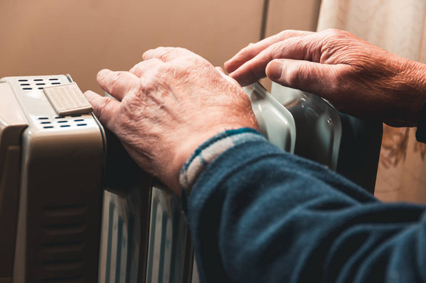 Un vieil homme se réchauffe les mains sur un radiateur électrique. En dehors de la saison, le chauffage central est retardé, de sorte que les gens doivent acheter des appareils de chauffage supplémentaires pour garder les maisons au chaud malgré l'augmentation des factures d'électricité
. - Photo, image