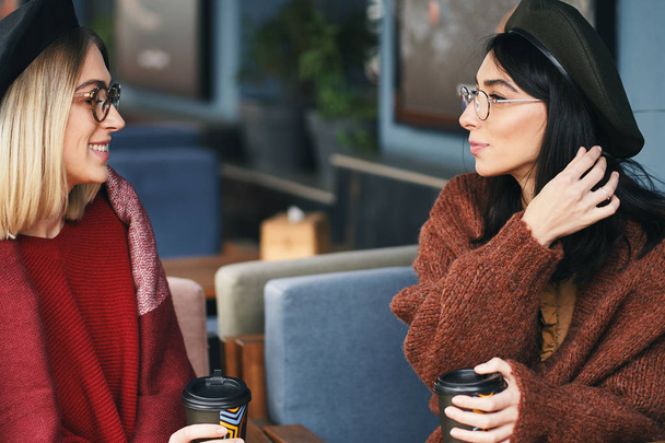Otoño invierno retrato de dos mujeres jóvenes en un café al aire libre, beber café para llevar, hablar. Fondo urbano. Hermanas.
 - Foto, Imagen