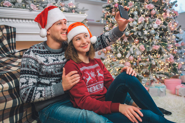 Delightful young couple sit together. They pose on camera. Man holds phone in hands and take selfie. They smile. Couple is in decorated room. - Valokuva, kuva