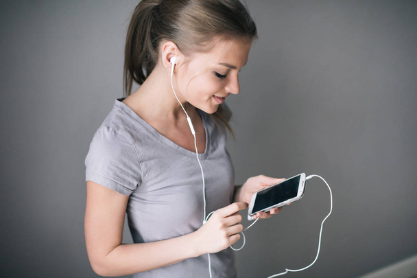 Young beautiful girl listening to music with headphones while showing blank screen mobile phone over gray background. - Fotografie, Obrázek