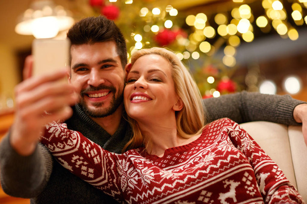 ευτυχισμένο ζευγάρι στο ζεστό πουλόβερ λαμβάνοντας αυτοπορτρέτα εικόνα με το smartphone στο σπίτι - Φωτογραφία, εικόνα