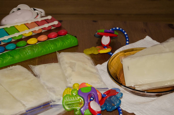 Latte materno congelato in sacchetti di vetro e sul tavolo di legno, circondato da giocattoli per dentizione e xilofono di mucca, latte materno pronto per essere conservato nel congelatore - Foto, immagini