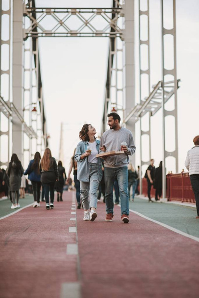 Young couple walking on the bridge with pizza and cold drinks - Фото, изображение
