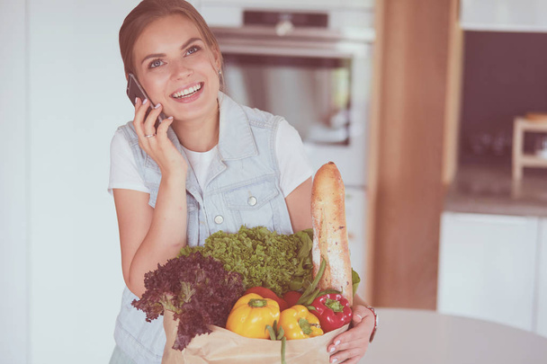 Smiling woman with mobile phone holding shopping bag in kitchen - Фото, изображение