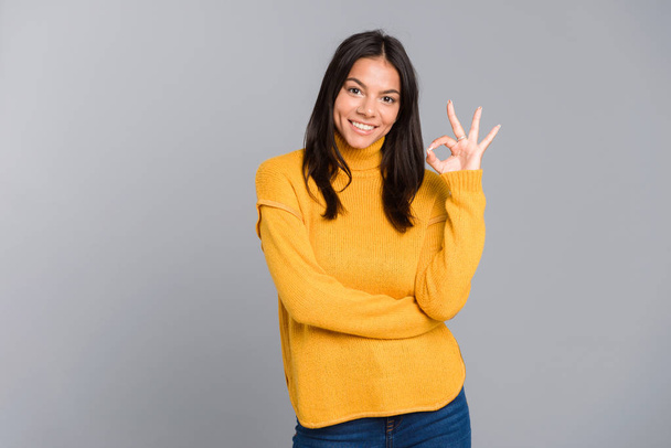 Portrait of a smiling young woman dressed in sweater standing isolated over grey background, showing peace - Fotoğraf, Görsel