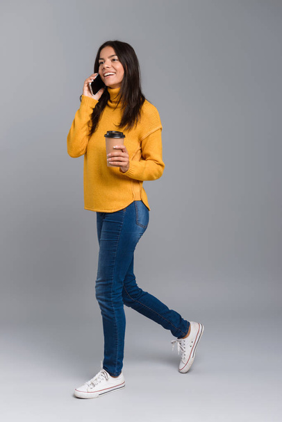 Full length portrait of a smiling casual young woman isolated over gray background, holding takeaway coffee,talking on mobile phone - Foto, Imagem