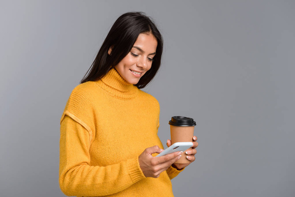 Portrait of a happy casual young woman isolated over gray background, holding takeaway coffee, using mobile phone - Фото, изображение