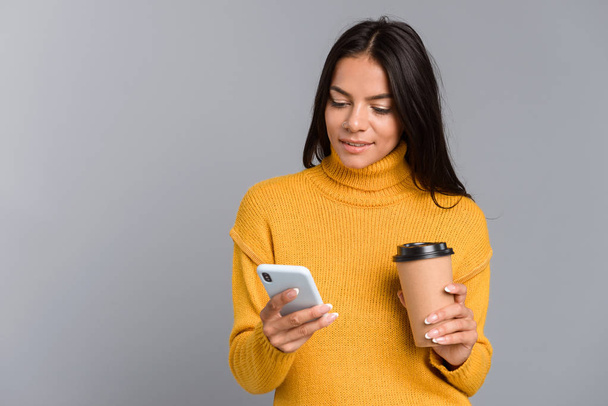 Portrait of a casual young woman isolated over gray background, holding takeaway coffee, using mobile phone - Foto, Imagem