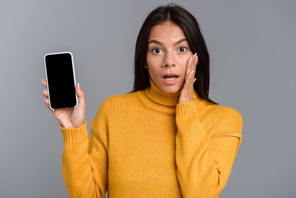 Portrait of a shocked casual young woman isolated over gray background, showing blank screen mobile phone - Valokuva, kuva