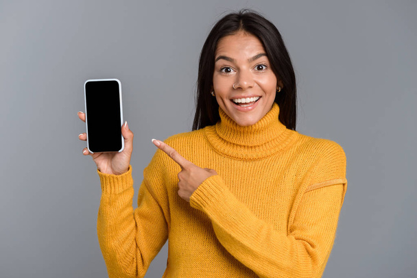 Portrait of a happy casual young woman isolated over gray background, showing blank screen mobile phone - 写真・画像