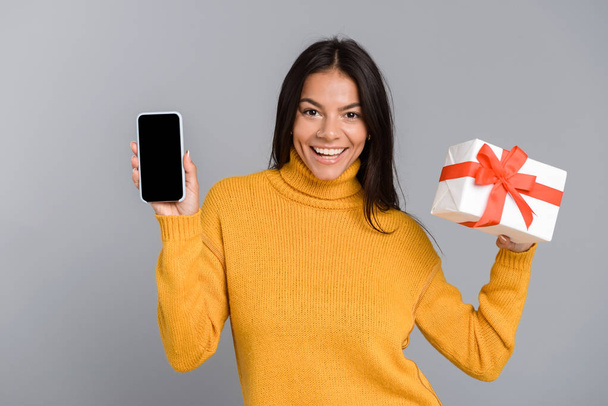 Portrait of a cheerful casual young woman isolated over gray background, showing blank screen mobile phone, holding present box - 写真・画像