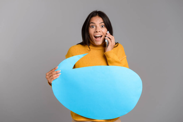 Portrait of an excited casual young woman isolated over gray background, talking on mobile phone, showing blank speech bubble - Photo, Image
