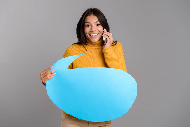 Portrait of a happy casual young woman isolated over gray background, talking on mobile phone, showing blank speech bubble - Foto, immagini