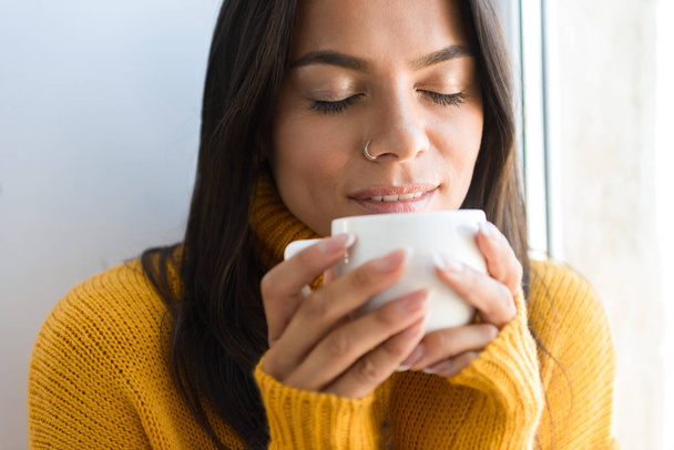 Primer plano retrato de una encantadora joven vestida con suéter sentada en la ventana en el interior, bebiendo una taza de té
 - Foto, imagen