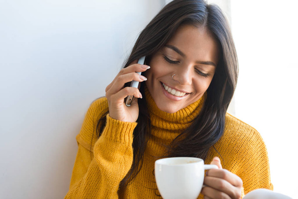 Close up portrait of a lovely young woman dressed in sweater sitting at the window indoors, holding cup of tea, talking on mobile phone - Photo, image