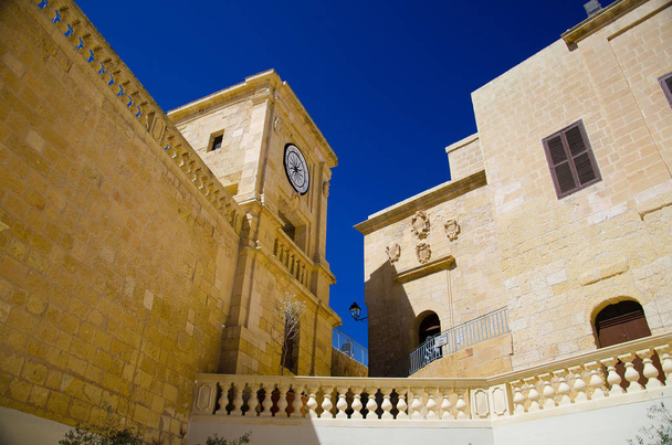 View of yellow buildings with watch in the old medieval Cittadella tower castle, also known as Citadel, Castello in the Victoria Rabat town, Gozo island, Malta - 写真・画像